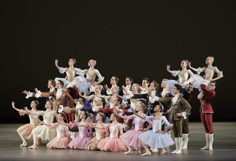 The cast of The Goldberg Variations knee and stand in a big formation. They gesture with their right hand. 