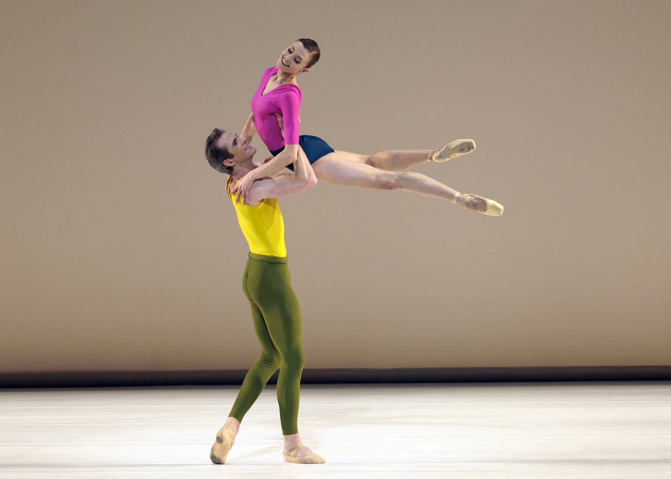a beautifully happy positive duet... a man in a bright yellow top and olive green tights swings his female partner in the air. she has bare legs pointe shoes, a bright pink top and navy blue short shorts.. This seems to be a neo classical square dance