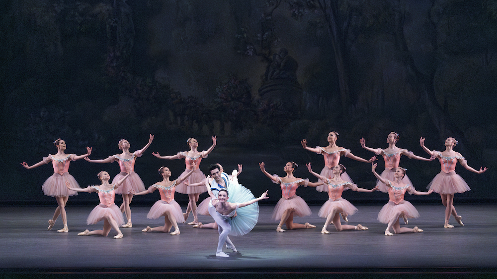 a chorus of the NYCB women in short pink tutus, bodices decorated with flowers and floral crown,pose upright or on one knee  as a male/female couple in blue tones smile at the audience... the male holds the femail suspended on his knee, her arms extend backward like wings 