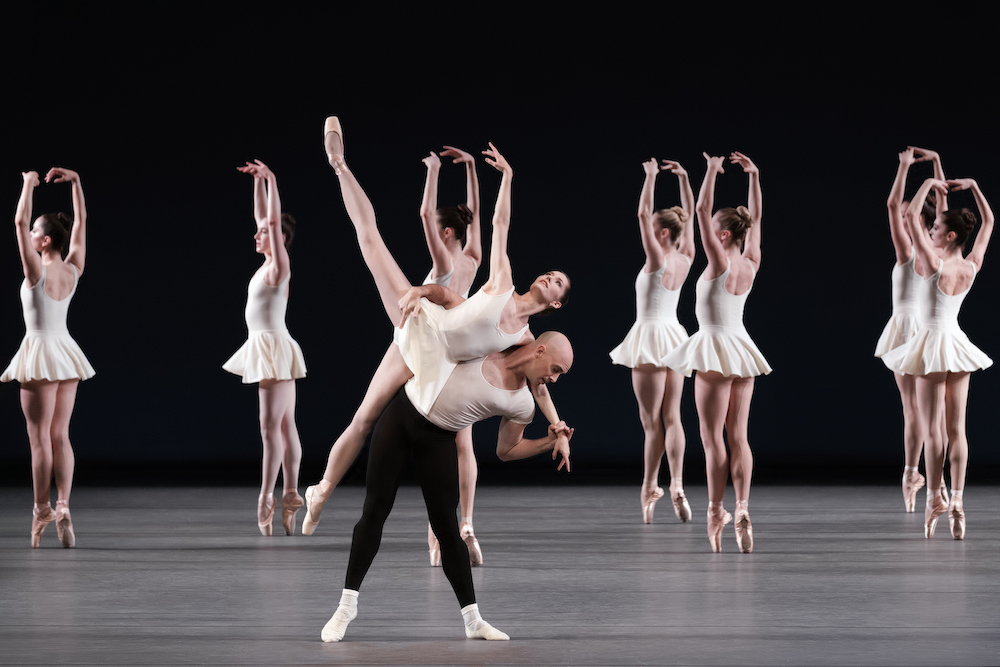 ballerinas in white short dresses face the back stage as a soloist similarly dressed perches atop the back of a man in a white t shirt with black tights 