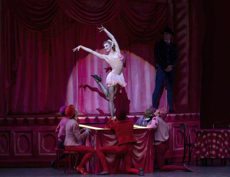 in a red almost cartoon like exaggerated set, a female dancer clothed in what looks like a fancy peach slip dress shimmies as men on a table below her oogle. On the stage with the shimming gal is a male figure in black who stands menacingly to the side, not brightly lit, but we can see he is a thug figure.