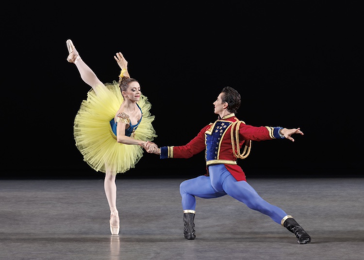 a woman in a bright yellow tutu with a blue bodice is supported as she leans forward on one leg in a penchee arabesque. Her partner dressed in a red suit jacket decorated with  blue and gold regalia, blue tights, and black boots, supports her from the position of a forward lunge. 