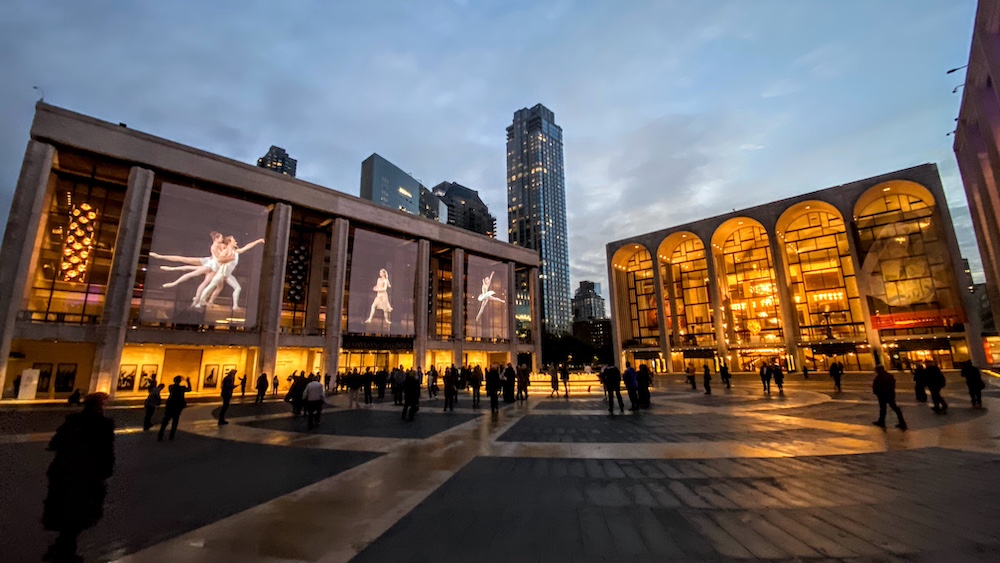 panoramic photo of Lincoln Center Plaza at Dusk. There are huge photos of scenes of larger than life NYCB dancers hanging at the balcony level of the Koch Theater