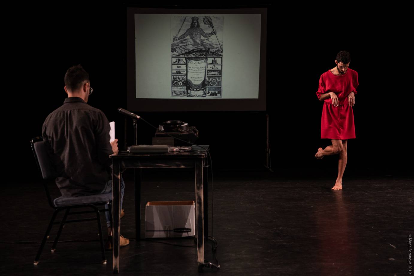 A man sits at a desk in front of a microphone while another man in red makes a prancing cat shape