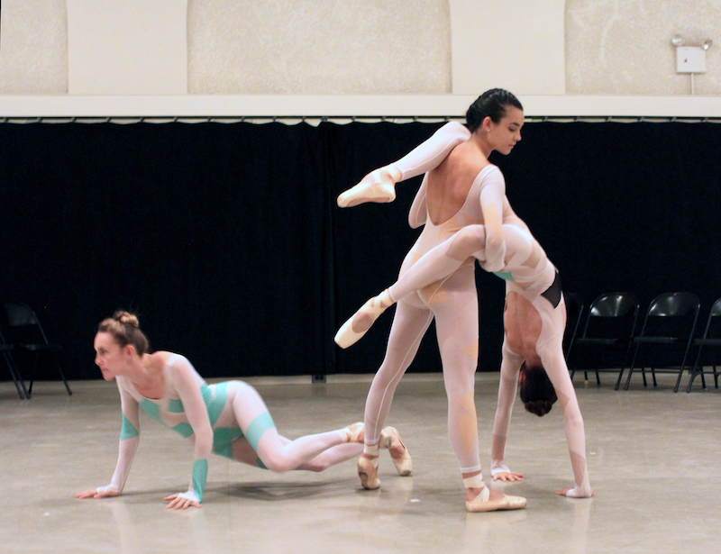 Three dancers in long-sleeved unitards. One dancer is on the floor with her arms and feet in pseudo plank position. A second dancer stands and supports the third dancer who is in a handstand position.