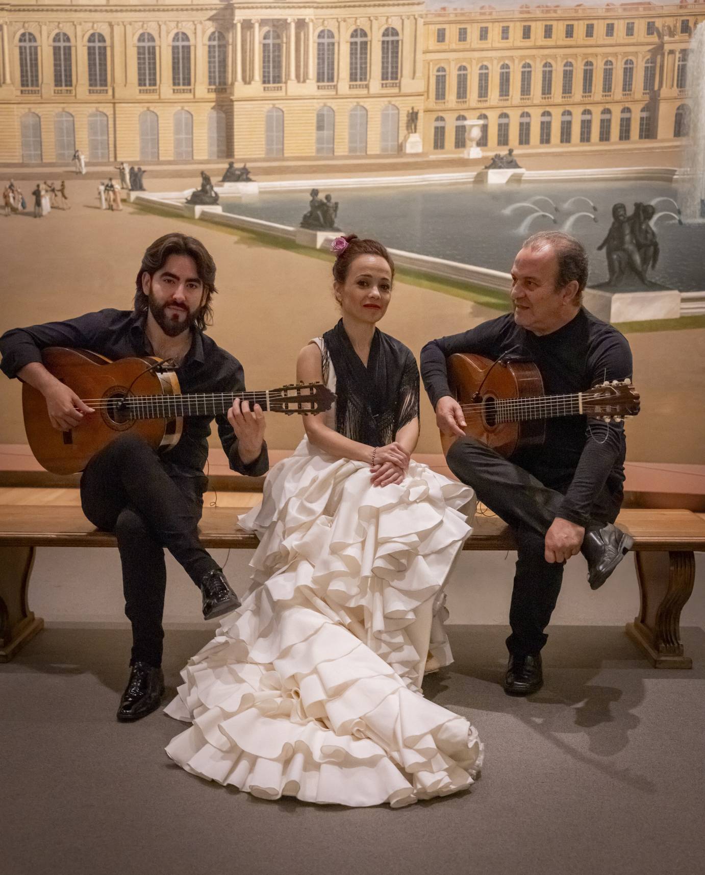 A woman in a white dress and black shawl is casually speaking with two musicians who flank her.