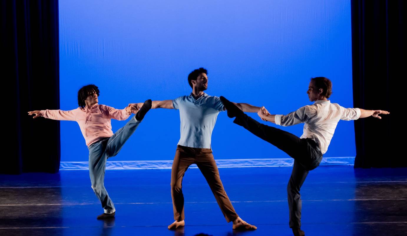 against a blue background three men, portraying a father and two sons hold on to each other. The faterh in the center seems proud and tall as the sons lean away from him mirroring one another with one leg lifted high in the air 