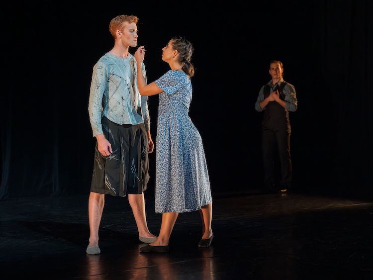 dressed in muted colors of blue and grey a young man stands staring at a pleading mother figure, behind them a man fades into the background, representing a missing father.