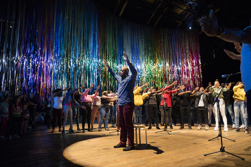 Vy Higginsen's Sing Harlem Choir stands in a semi circle around a male soloist. They all have their arms raised.