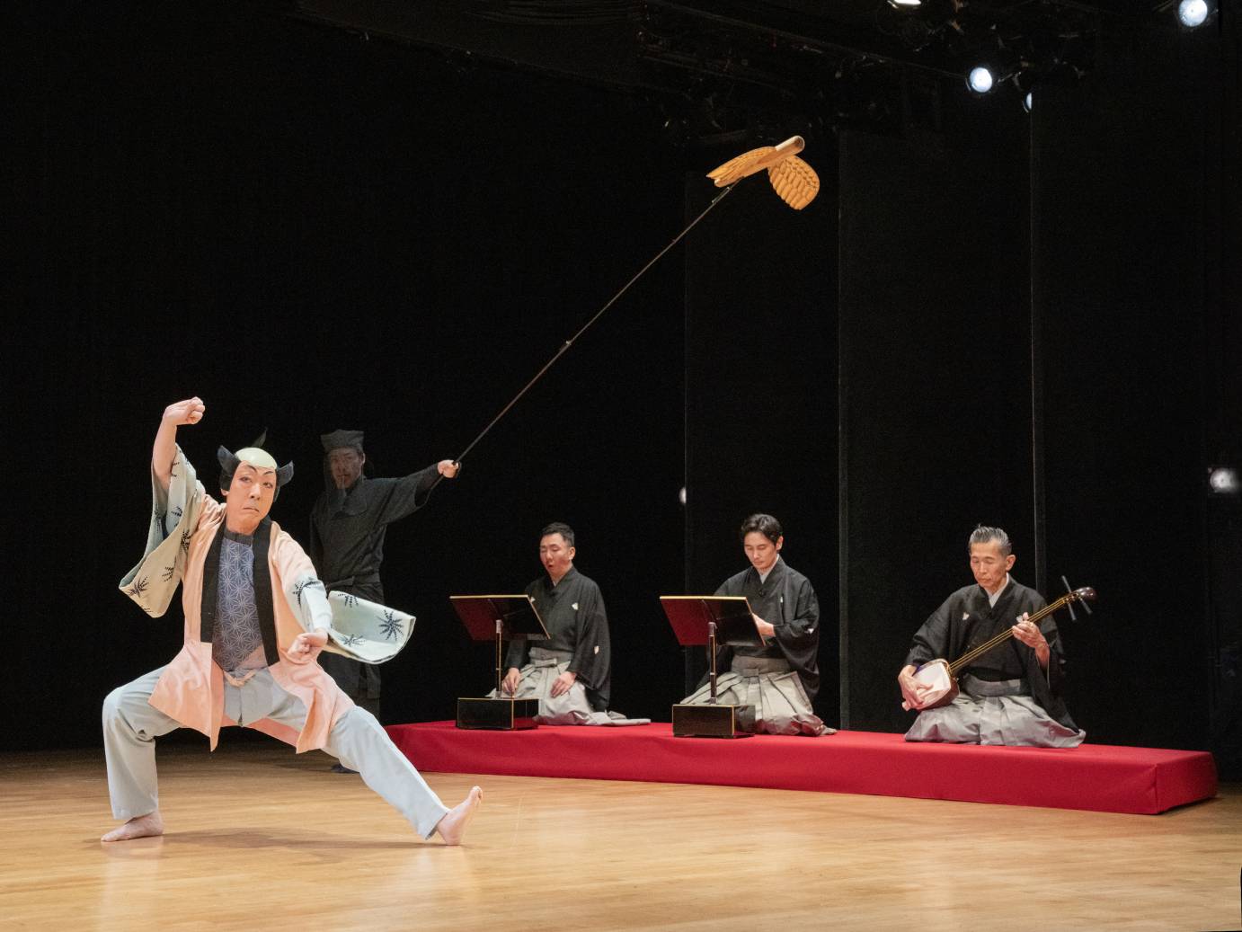 Male dancer wearing stylized black hair, with a bald spot on top, define makeup and short kimono, extends and flexes his right leg and foot. His right arm is above his head. In the background, sit three musicians, with legs folded beneath, on a platform covered with red cloth