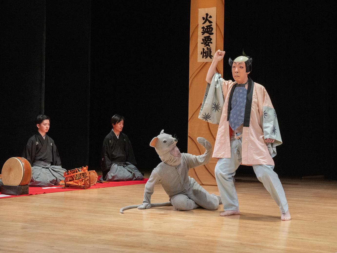 The servant character stands above the human sized mouse character who is beseeching from the floor. Two musicians sit with knees folded beneath their bodies looking ahead.