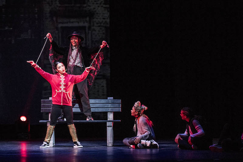 A woman standing on a bench manipulates strings that are attached to another dancer's wrists like a marionette. 