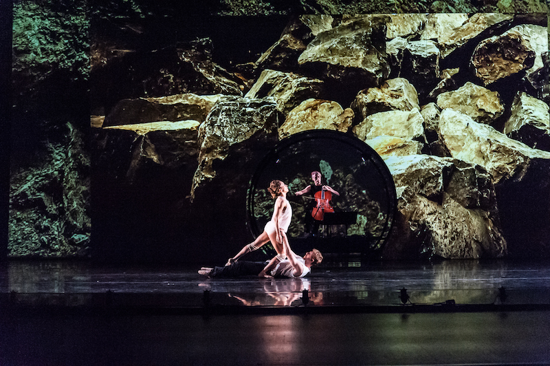 A man lies on his back as he support the weight of his partner who is suspended above him. Cellist Zoe Keating plays behind them. A larger-than-life project of boulders play behind the trio.