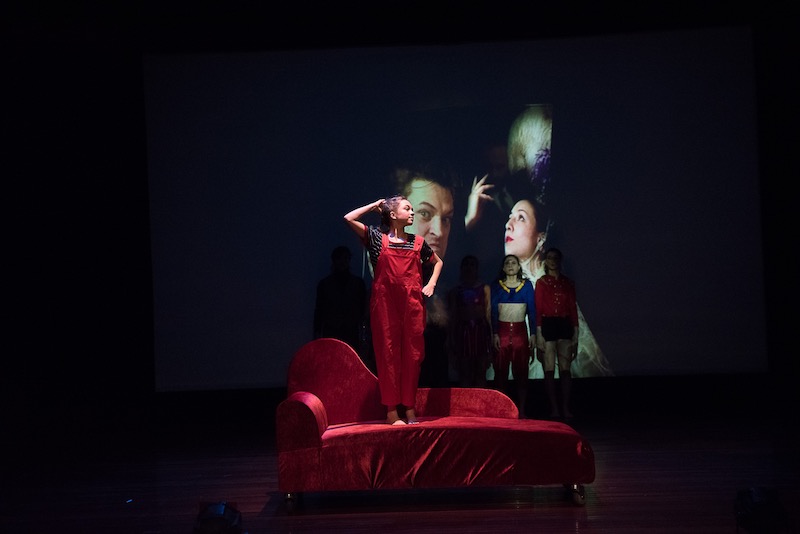 A young girl in red overalls stands on a red velvet couch. Other dancers stand and watch her in the background.