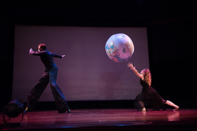 A man walks on stilts while a woman in the black dress hits a beach ball that has a map of the world printed on it.