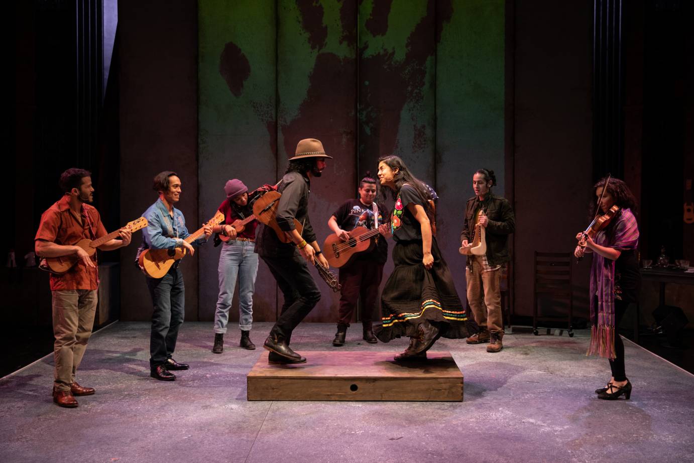 A man and a woman stomp their feet on a platform, surrounded a group of strumming musicians