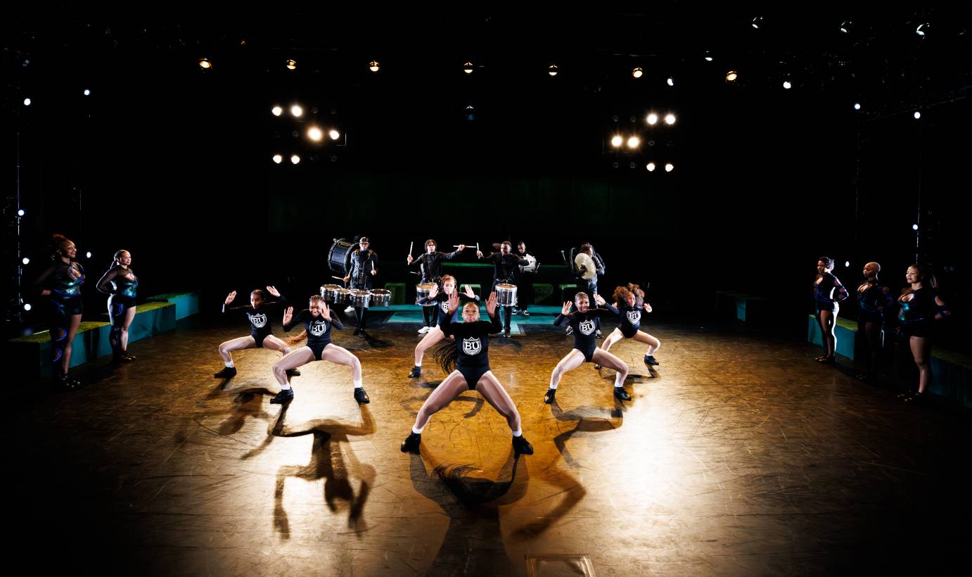 Boys upstage drumming behind a phalanx of girls in second position facing the audience.