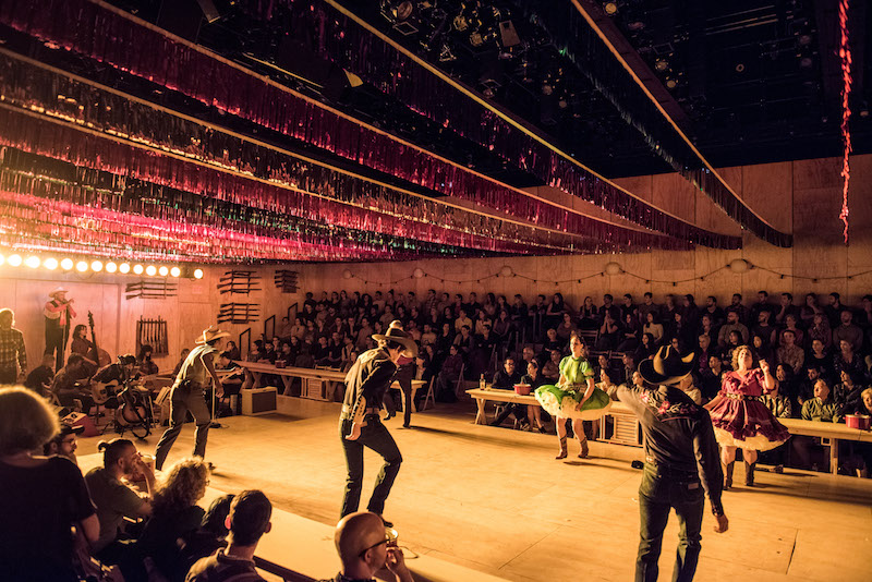 A shot of the stage and audience of Oklahoma at St. Ann's Warehouse. Performers, some in cowboy hats and boots, dance on stage. Colorful streamers are hung above them. Audience members are seated on two sides of the stage.