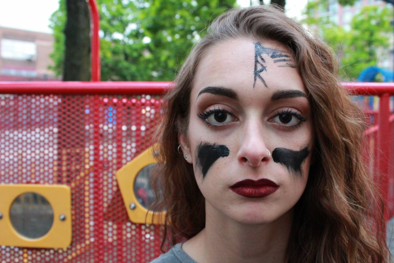 A girl with red lips and black streaks on her cheeks and forehead poses in front of a playground