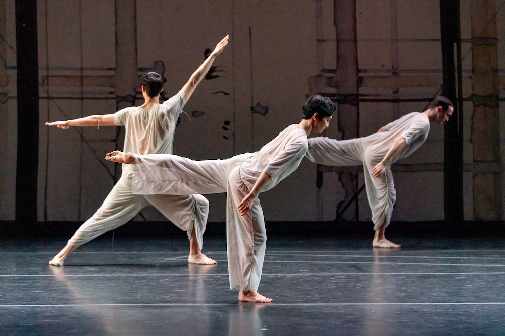 a trio of dancers move against the artistic background consisting of lines and brush strokes. beautifully lit wearing sheer blue-grey flowing jump suits, two dancers lean into a meditative arabesque while behind them another dancer faces the backstage in a lung one arm extended upward the other to the side