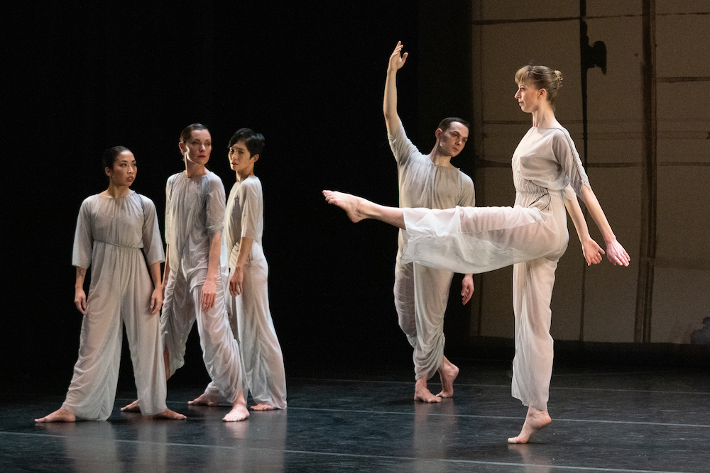 5 dancers, grouped as a trio, and two soloists, wear flowing sheer light blue-grey pant suits. some with sleeves some not. the trio peers at a woman with an erect spine kicking her leg forward, a man, bheind the woman soloist  poses, one leg  bent slightly behind him and one arm in a high upright position