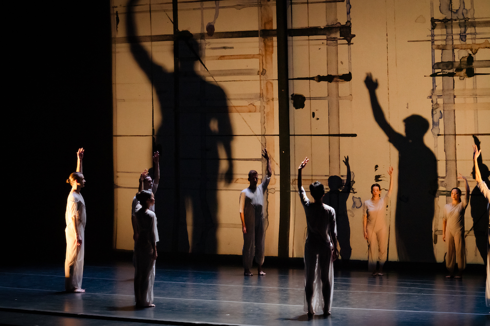 the Pam Tanowitz company on stage 10 dancers against an artistic background of brown and black brush strokes stand erect, each with one arm up in the air. they are lit dimly and their shadows loom above behind them, giants in the background