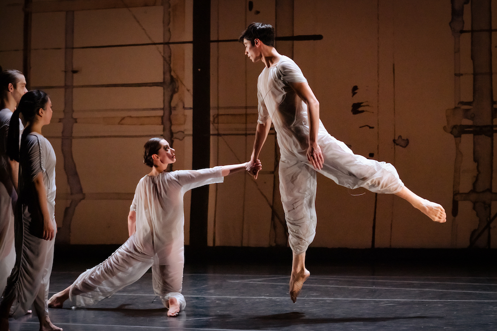 a woman dancer kneeling on her left knee, with her rlight leg extended on the floor heolds the hand of a man who is at the height of an arabesque jumb, tow other dancers look on 