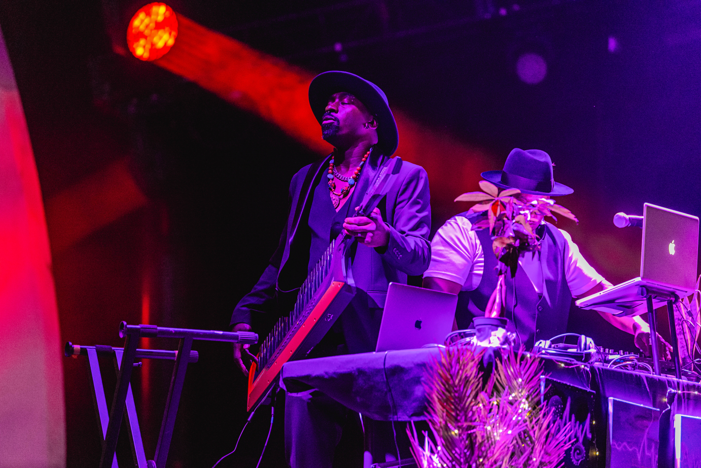 A man in a fedora and suit jacket plays guitar, his eyes closed. To his side, a man in a hat works DJ equipment. 