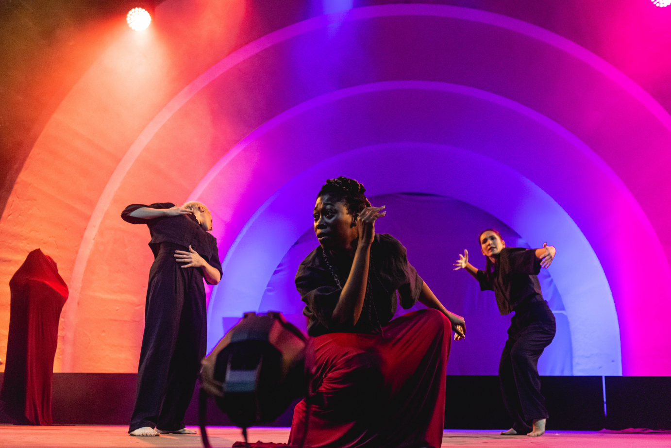 Against arches of purple, pink, and orange light, the members of Passion Fruit Dance Company groove. Downstage, one kneels while, behind her, two gesticulate. 