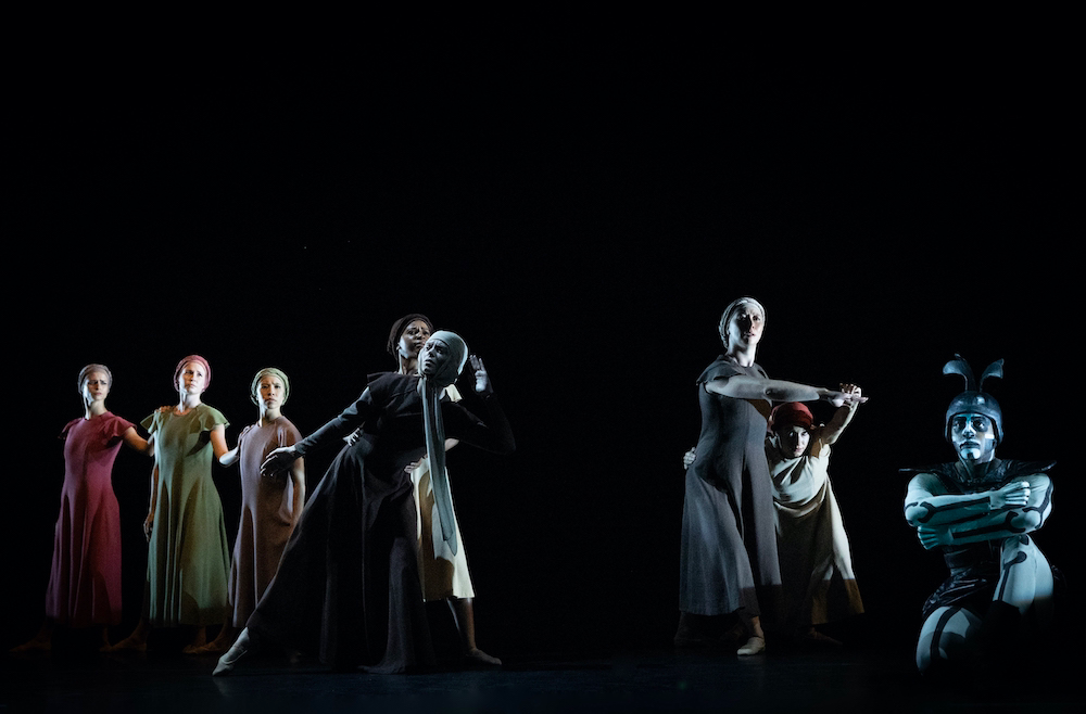 a group of somberly clothed women dressed as refugees look out into the distance with a figure representing death seated in front of them 