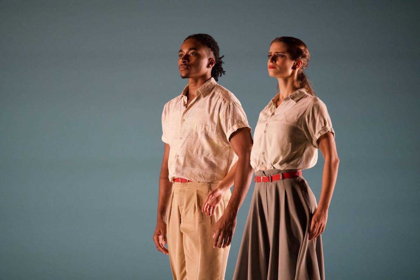 A couple, a Black man and a fair-skinned woman, in cacky colored clothes, stand still and poised, as if looking out towards the future. 