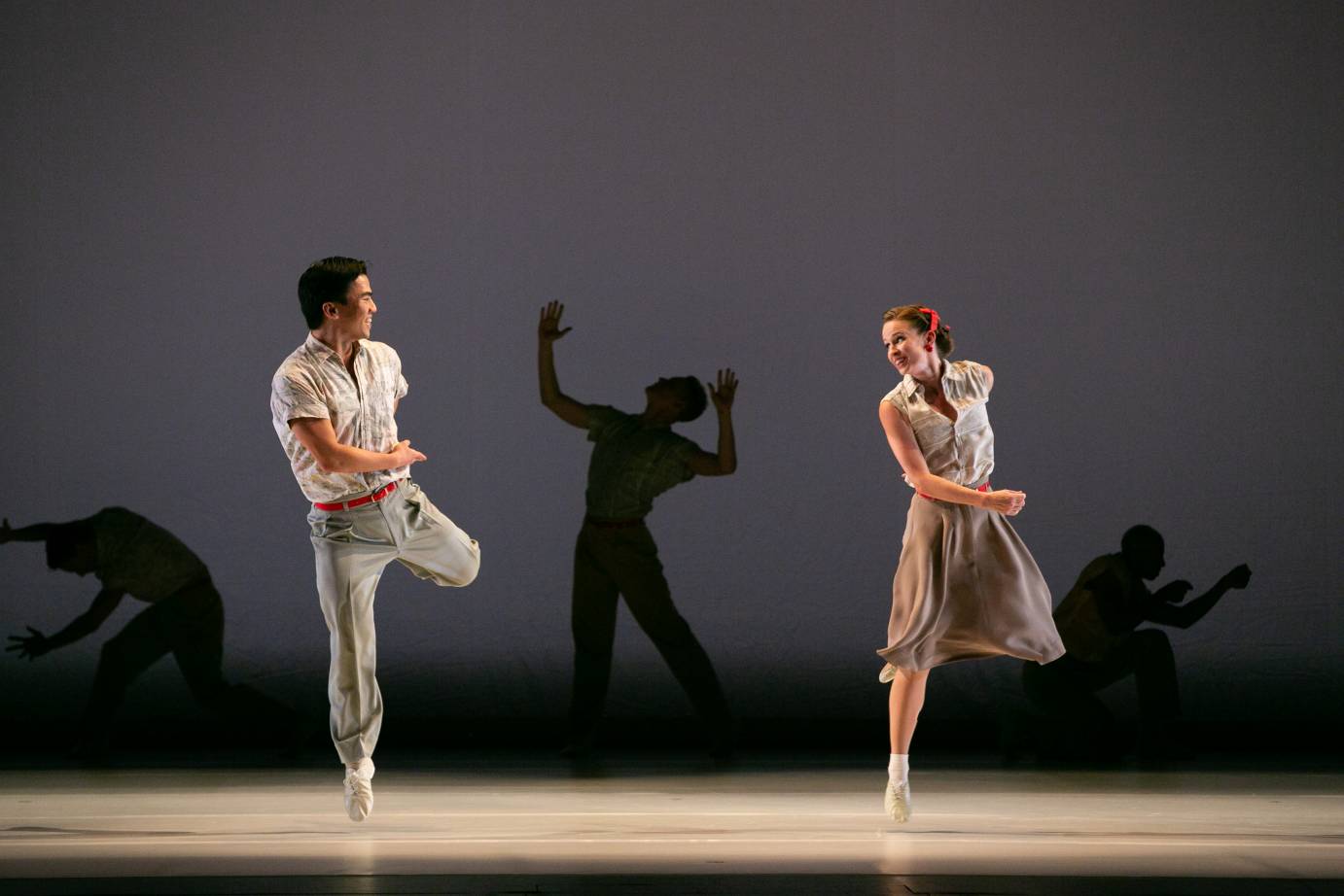 a spritely couple, a male and female in 1940ish clothes, dance together,mirroring eachother. Each stands on one leg with the other lifted up behind them... there are other dancers silouhetted in the back ground..