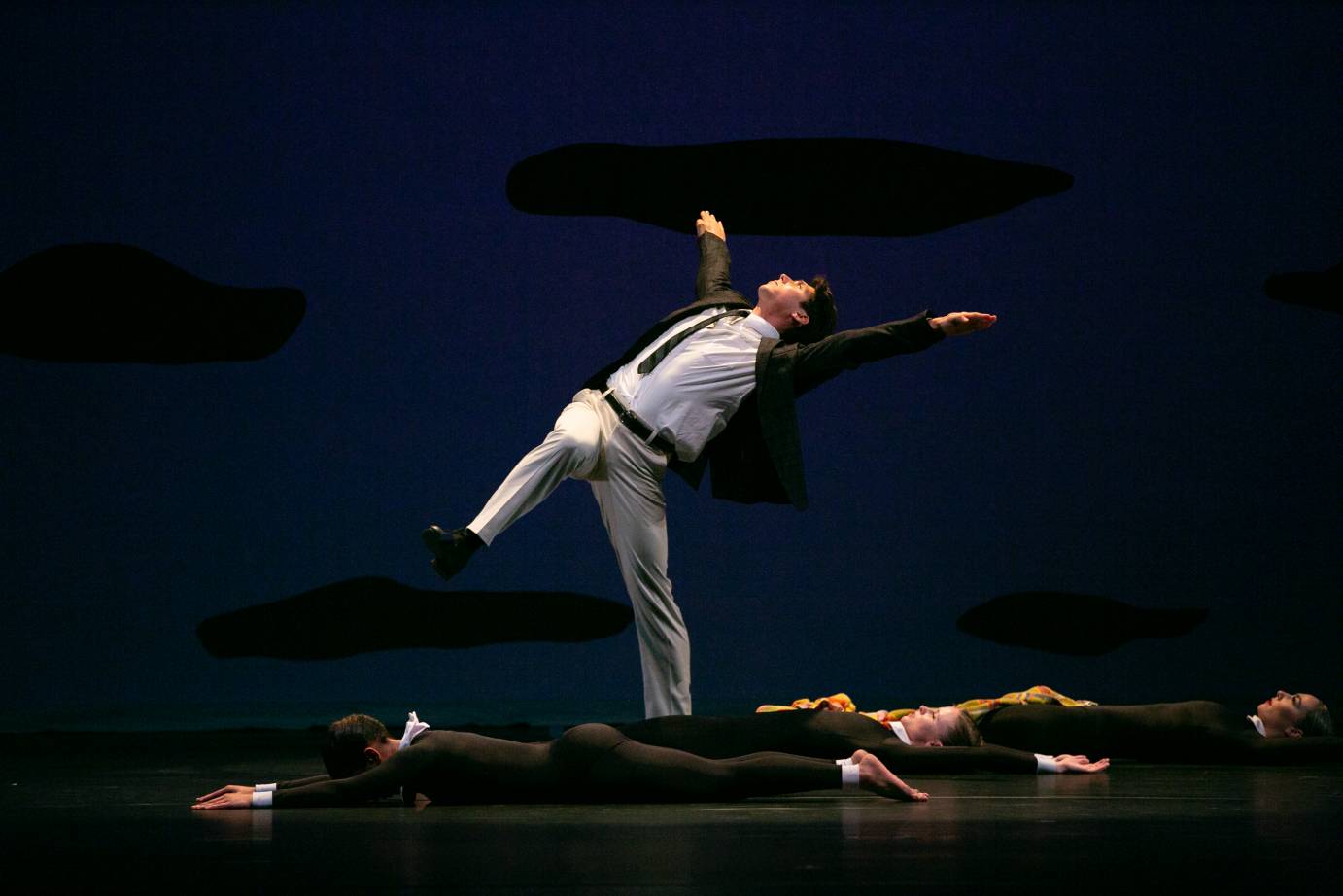 amidst a litter of possibly dead bodies a young man in cacky pants and a sportscoat spreads his arms wide while arching his back and looking upwards, standing on one leg