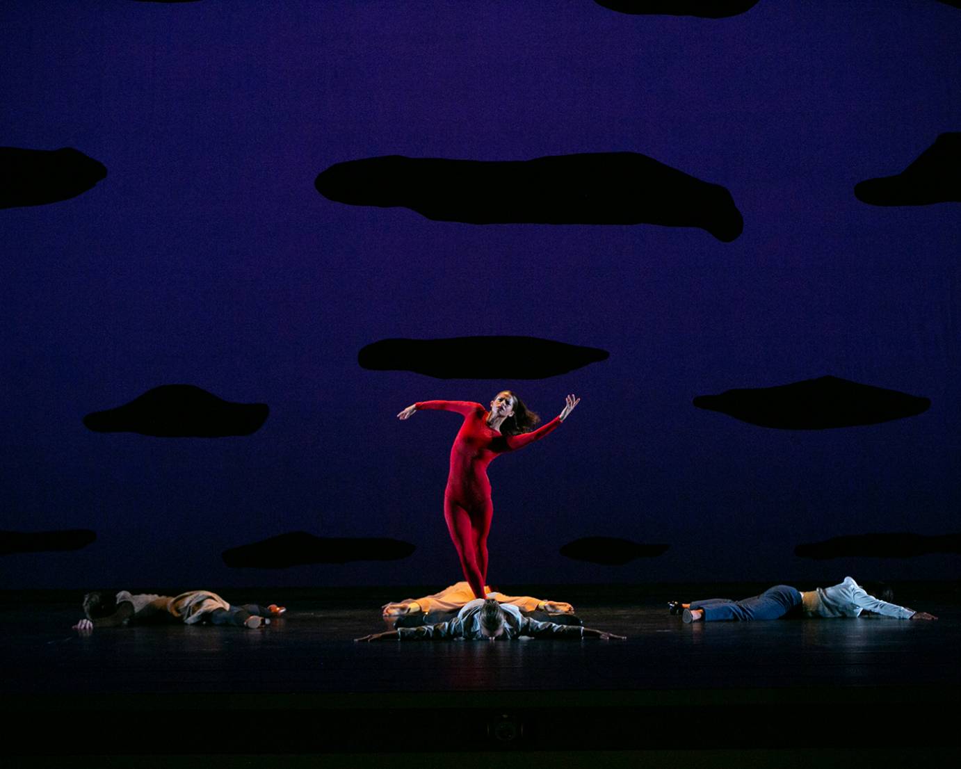 woman in red unitard stands in the center of a group ofpeople lying down on the floor,her arms are curved she leans slightly to her left and her hair flows