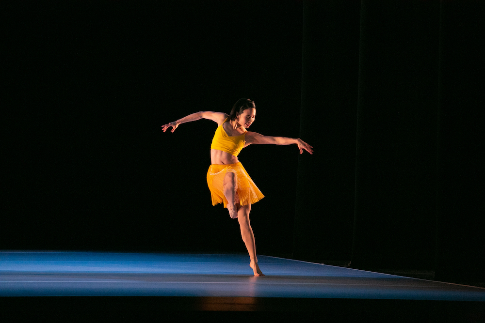 a woman in yellow, a yellow top with bare midriff and a pleated yellow mini balances on one foot in a pool of light