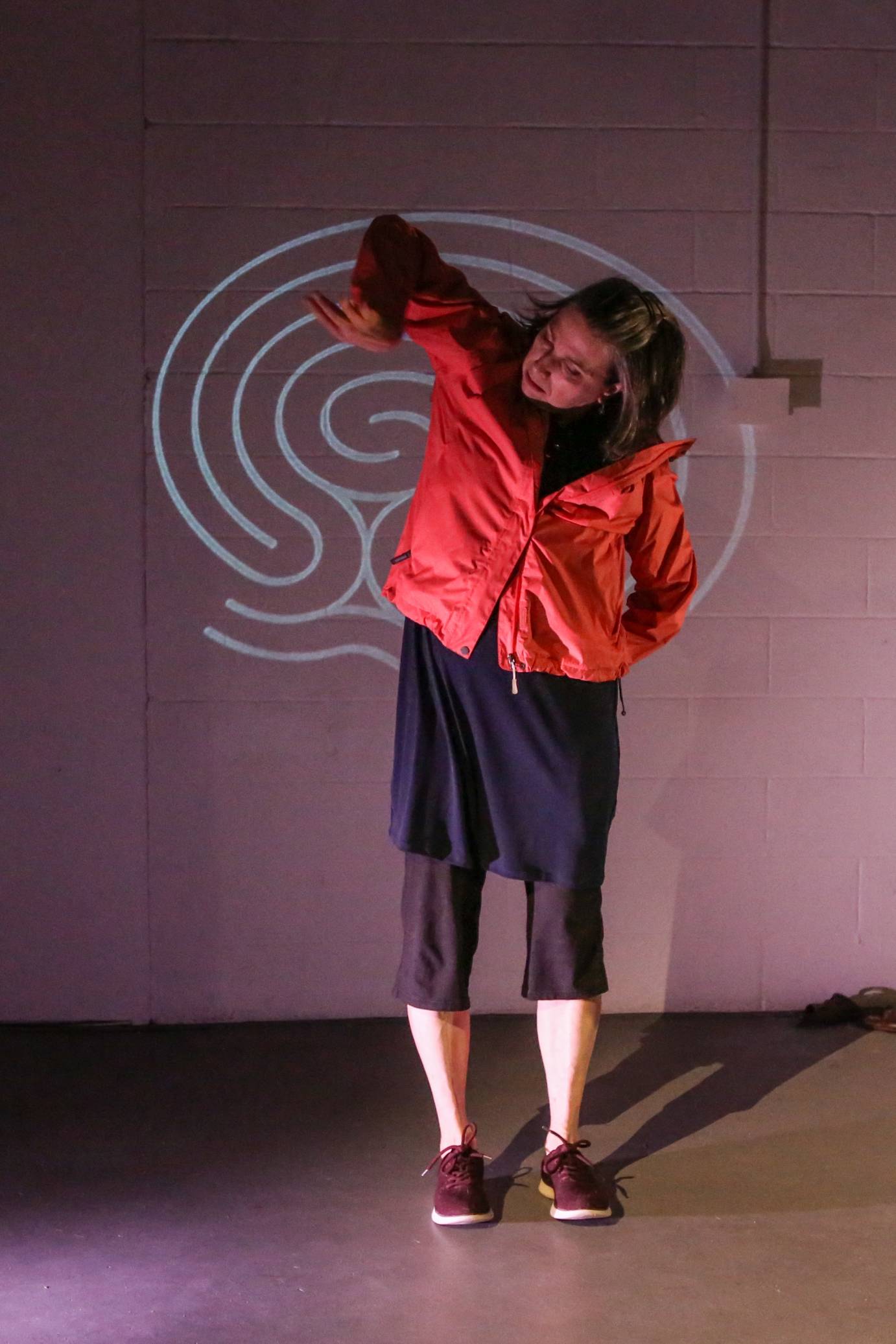 White woman with chin length brown hair dressed in a red dress and black pants right elbow lifted with hand hanging down in front of a white spiral projection on a tan wall