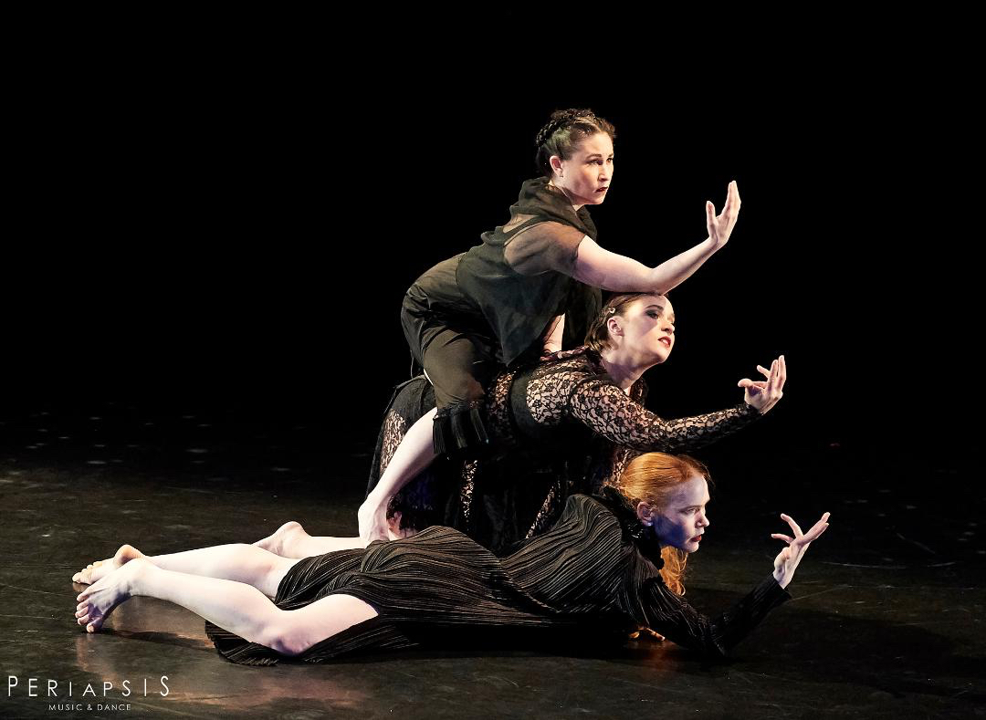 Three women lie stacked on top of one another, one arm extended beseechingly