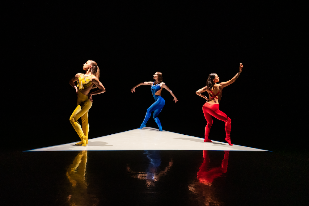 a triangle of light encases three female dancers dressed in bright tight  primary  colors