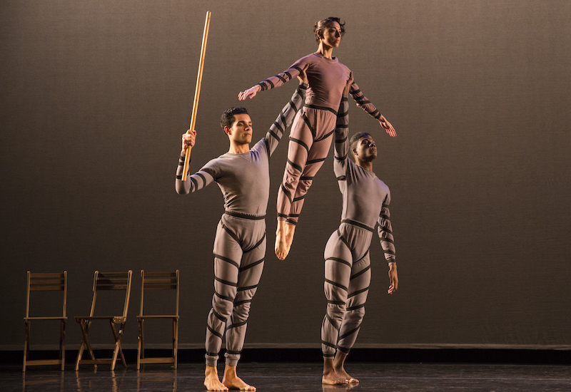 Two dancers lift a dancer between them over their head. Three wooden chairs are in the background. One dancer holds a wooden staff.