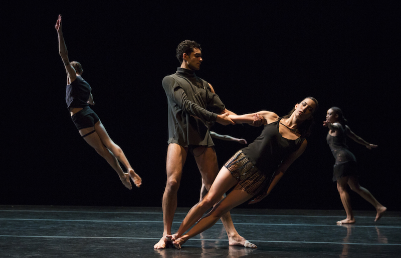 In the foreground, one dancer leans and is held by the arm by another artist. A dancer, with their back facing the audience leaps high into the air. 