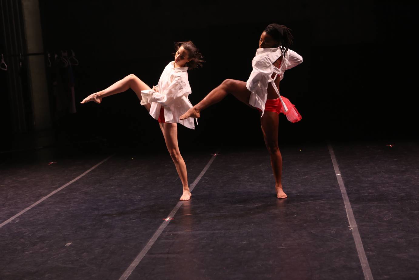 two women in long white shirts and red short red pants with a tall of netted tulle kick up their leg in unison