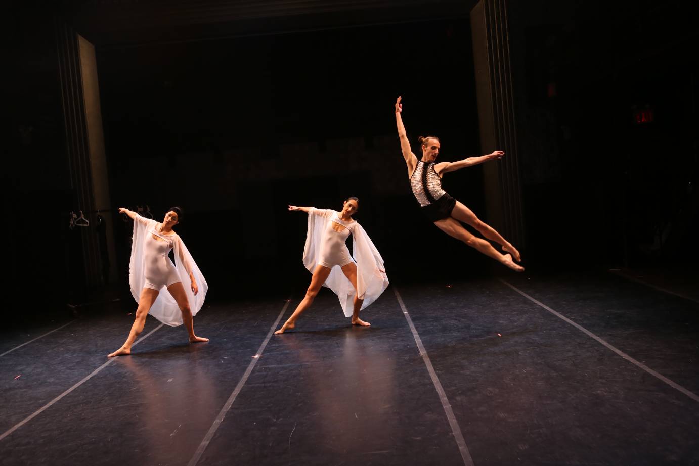 two women resembling angels in short white unitards with flowing white shawls stand with legs wide apart and stare outwards as a male dancer in shorts flies high above them in the crest of a leap 