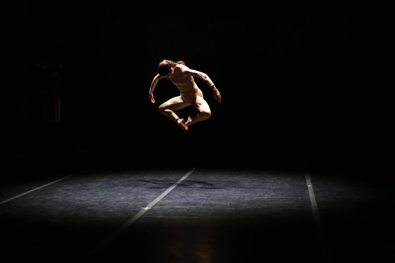 Jaqlin Medlock in a light colored unitard hovers in mid air,her toes touching and the shape of her legs in the air resemble a diamond 