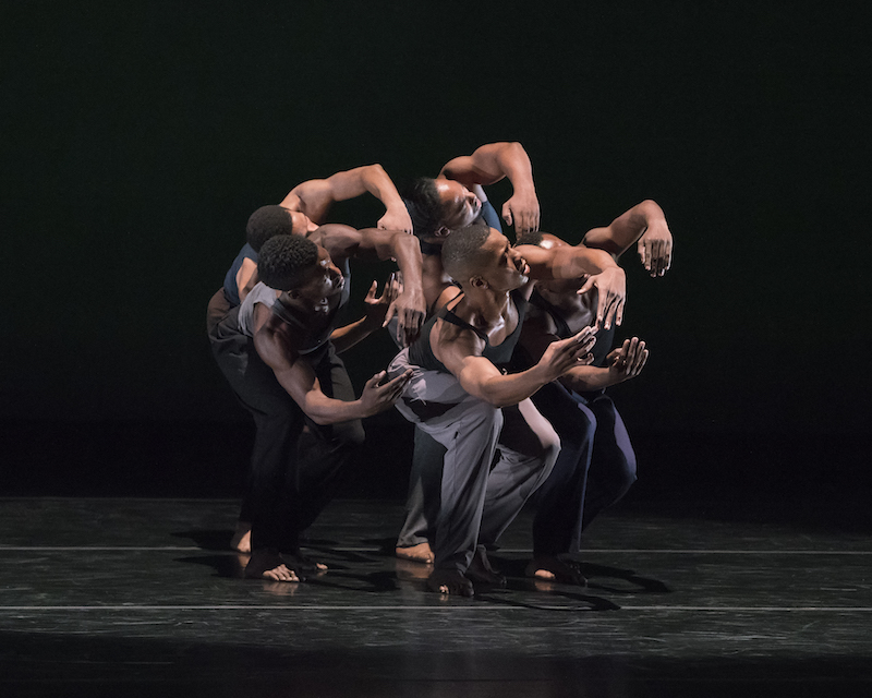 A group of men in slacks and tank tops curve their bodies to one side making a C shape with their arms