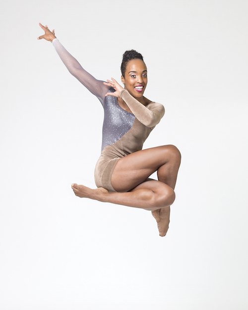 A dancer in a shimmery unitard smiles at the camera as she tucks her legs in a jump.