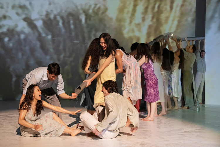 against a projected tropical background the company of dancers women in gowns, men in everyday khaki colored pants and oxford shirts play hold up a flexible water pipe that shoots water out on one of the female dancers who sits on the floor and laughts