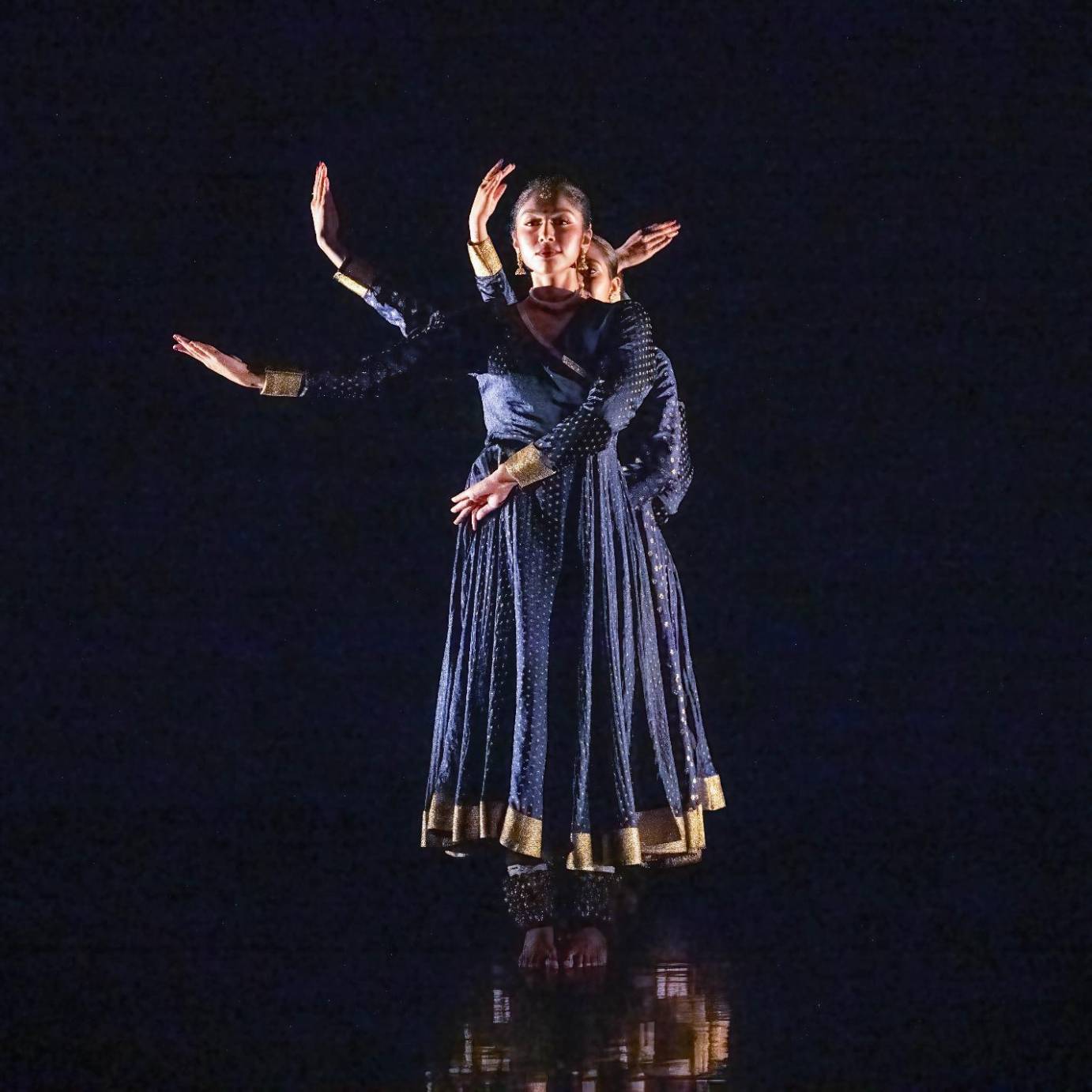 A line of women from downstage to up in long navy dresses look out to the audience in front of a black background, their arms bent and lifted in relation to the front woman like a hydra.