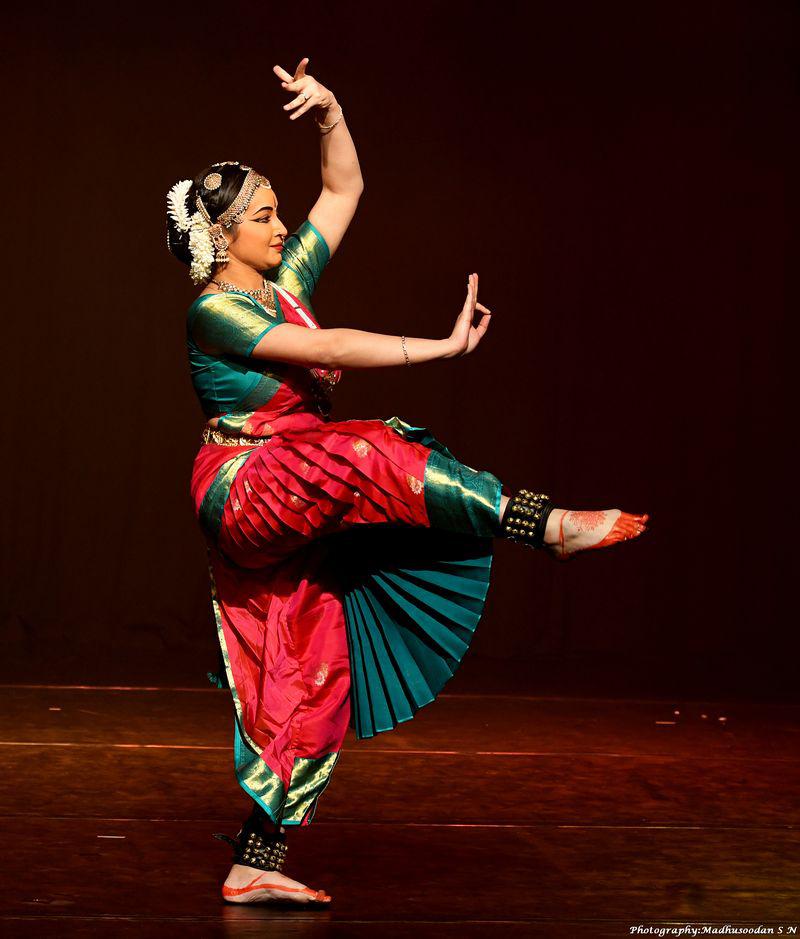 A woman dressed in green and red stands in profile pressing one arm with a flexed palm in front of her and the other arm, fingers open, float above her head. Her right leg is lifted, knee bent. 