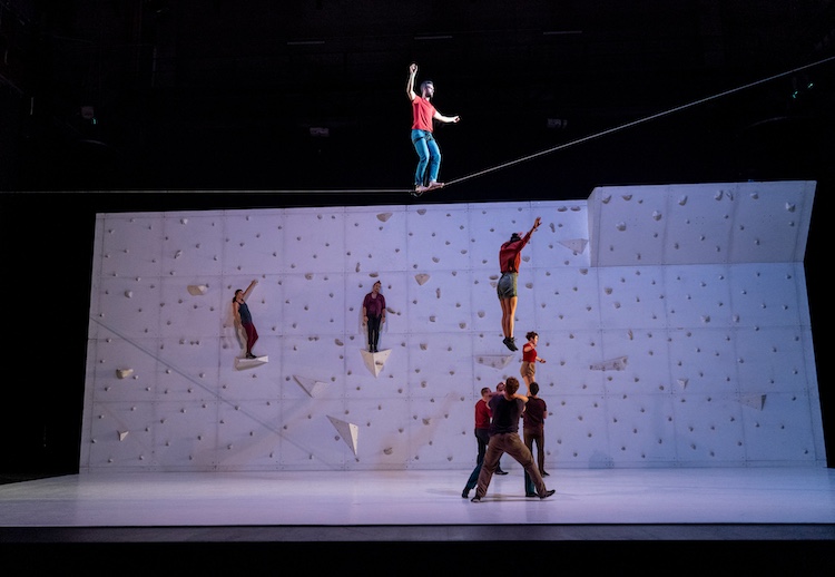 a man on a hire wire balances high above a company of dancers standing on a rock wall and on the stage floor. One dancer jumps high in the air under the tightrope walker.