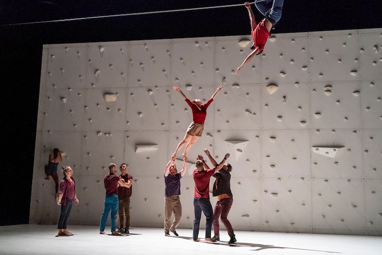 a dancer held aloft  by her feet reaches to a man hanging upside down from his knees on a tightrope abover her ...other cast mates look on 